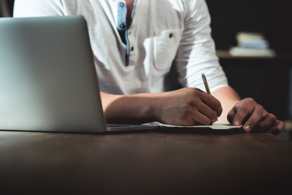 Cropped Shot Of Man Writing In Notebook During Work While Sitting At The Table With Laptop